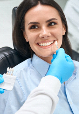 Woman in dental chair smiling as dentist holds shade guide to teeth